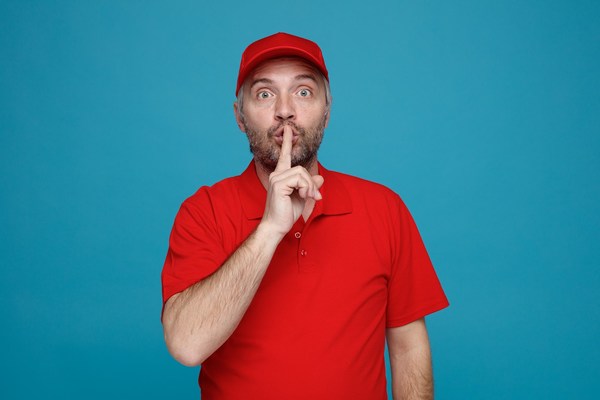 delivery man employee in red cap blank tshirt uniform looking at camera surprised making silence gesture with finger on lips standing over blue background